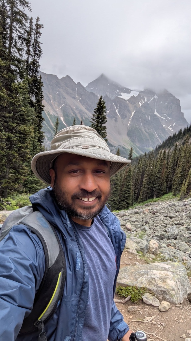 Steve in a sun hat and rain jacket on a rocky trail below cloud-capped peaks